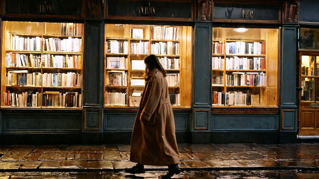 A finished environmental wide shot — a figure walking past lit shop windows on a wet street, warm reflections, atmospheric fog, the place as much the subject as the person.